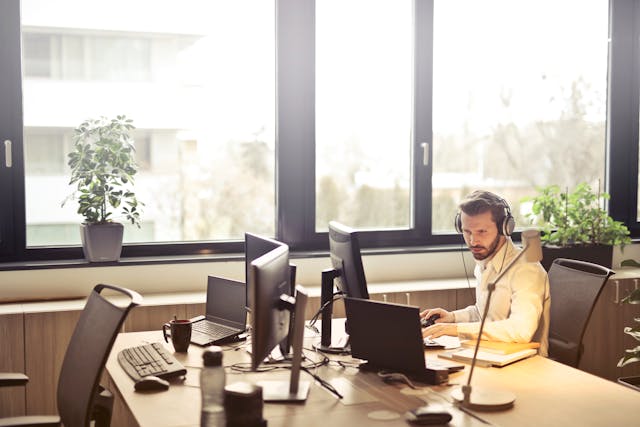 Karyawan menggunakan headphone sedang bekerja dengan beberapa monitor komputer di ruang kantor yang terang