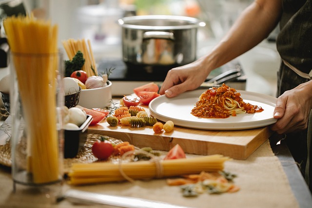 Seorang koki sedang menyajikan hidangan pasta dengan saus merah di dapur yang dilengkapi berbagai bahan makanan seperti pasta dan tomat